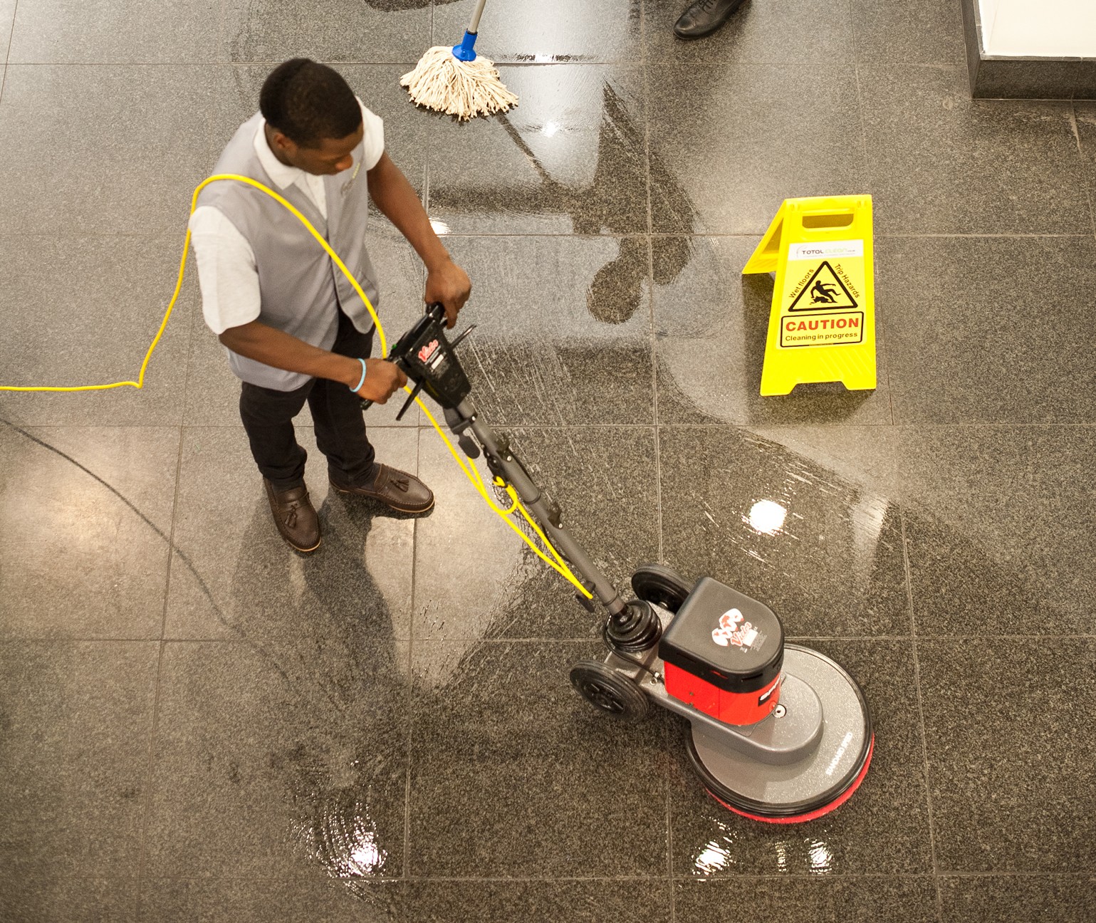 Woman deep cleaning commercial office window while a man hoovers in the hallway of the office